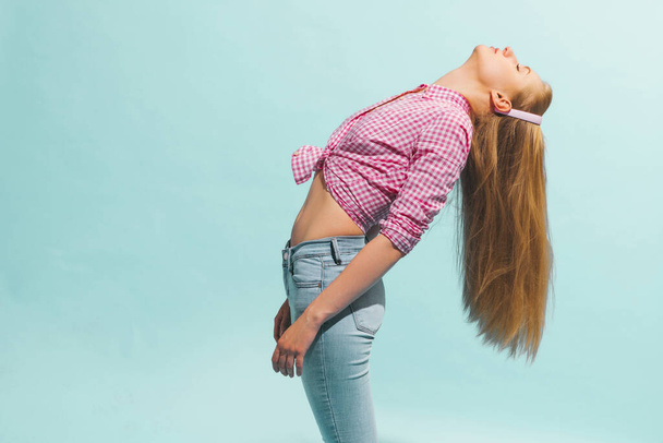 Model throwing your head back. Young beautiful girl with long blond hair wearing plaid shirt posing isolated on blue background. Concept of human emotions, facial expression, youth, aspiration, ad - Foto, imagen