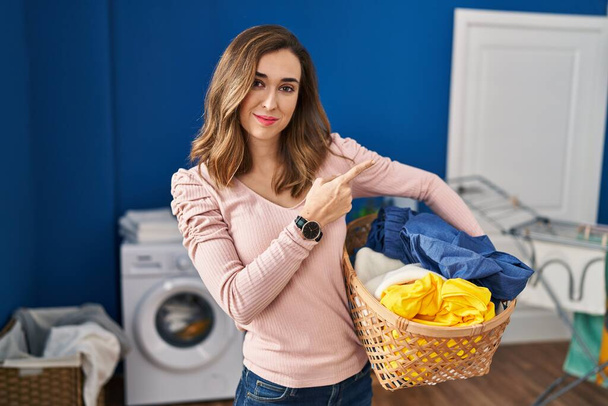 Young woman holding laundry basket pointing with hand finger to the side showing advertisement, serious and calm face  - 写真・画像