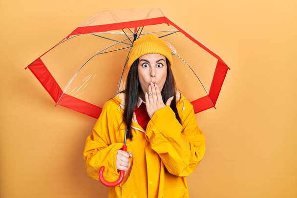 Jovem hispânica vestindo capa de chuva amarela segurando guarda-chuva cobrindo a boca com a mão, chocado e com medo de erro. expressão surpreendida  - Foto, Imagem