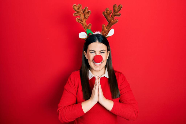 Young hispanic woman wearing deer christmas hat and red nose praying with hands together asking for forgiveness smiling confident.  - Photo, Image