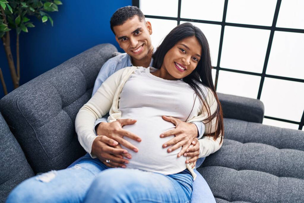 Jovem casal latino esperando bebê tocando barriga deitada no sofá em casa - Foto, Imagem
