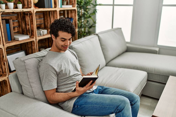 Young hispanic man reading book sitting on the sofa at home. - Fotó, kép