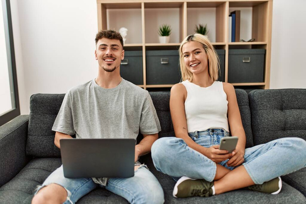 Jeune couple caucasien souriant heureux en utilisant un ordinateur portable et smartphone assis sur le canapé à la maison. - Photo, image