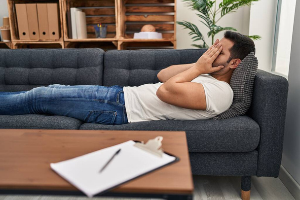 Hombre hispano joven estresado teniendo terapia mental tumbado en sofá en el centro de psicología - Foto, Imagen