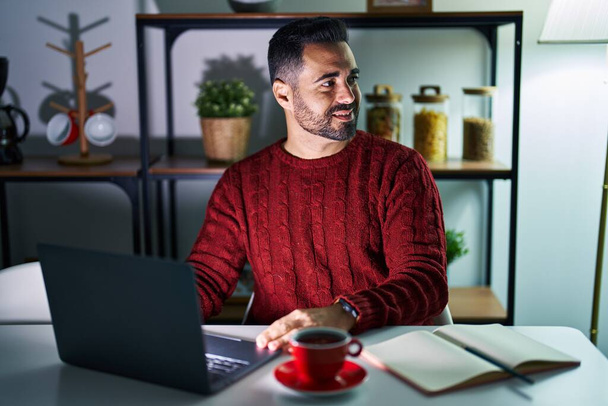 Joven hombre hispano con barba usando computadora portátil por la noche en casa mirando al costado, pose de perfil relajado con rostro natural y sonrisa confiada.  - Foto, imagen
