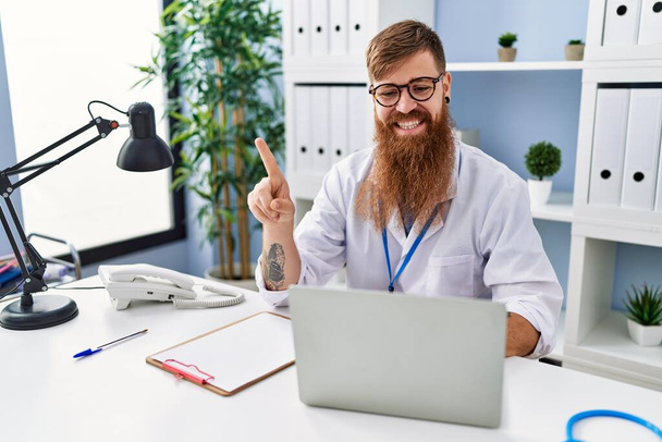 Redhead man with long beard wearing doctor uniform working using computer laptop smiling happy pointing with hand and finger to the side  - Zdjęcie, obraz