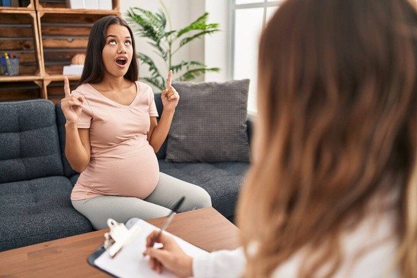 Young pregnant woman at therapy session amazed and surprised looking up and pointing with fingers and raised arms.  - Photo, image
