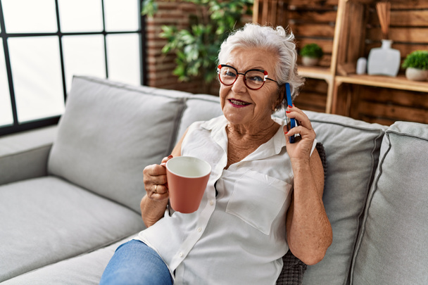 Starsza siwowłosa kobieta pije kawę i rozmawia na smartfonie siedząc na kanapie w domu - Zdjęcie, obraz