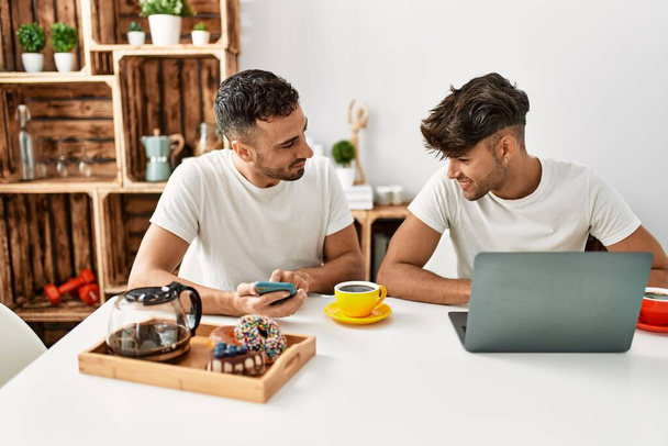 Δύο ισπανόφωνοι άνδρες ζευγάρι που παίρνουν πρωινό χρησιμοποιώντας smartphone και laptop στο σπίτι - Φωτογραφία, εικόνα