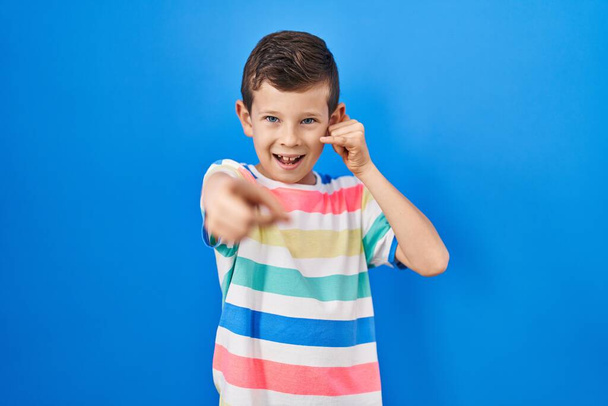 Jeune enfant caucasien debout sur fond bleu souriant faire parler au téléphone geste et vous pointant du doigt. Appelle-moi.  - Photo, image