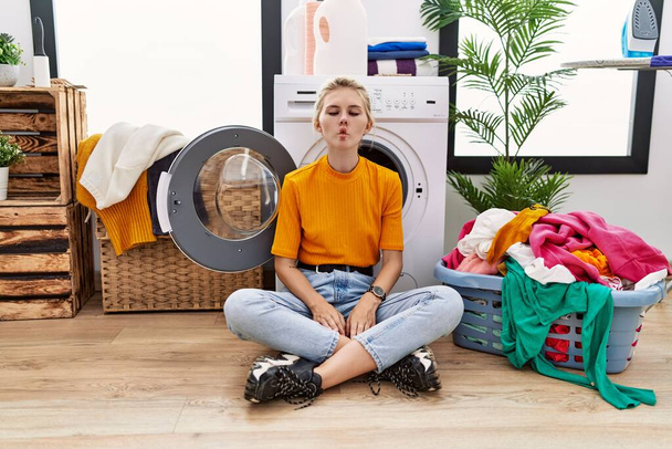 Young blonde woman doing laundry sitting by washing machine making fish face with lips, crazy and comical gesture. funny expression.  - Foto, afbeelding