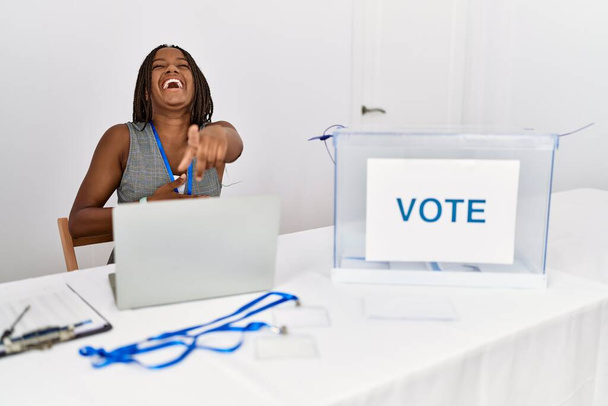 Jonge Afro-Amerikaanse vrouw die bij de politieke verkiezingen werkt... zit je uit te lachen, wijzend met de vinger naar de camera... met de hand over het lichaam, schaamte uitdrukking.  - Foto, afbeelding