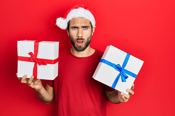 Joven hombre hispano vistiendo sombrero de navidad y sosteniendo regalos asustado y sorprendido con la expresión sorpresa y asombrado, miedo y cara emocionada.  - Foto, Imagen