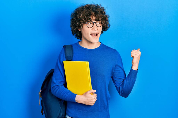 Bonito jovem vestindo mochila estudantil e segurando livro apontando polegar até o lado sorrindo feliz com a boca aberta  - Foto, Imagem