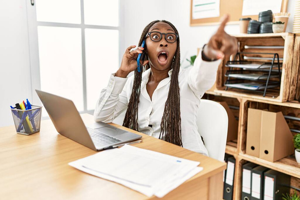Schwarze Frau mit Zöpfen, die im Büro arbeitet, telefoniert und zeigt mit dem Finger überrascht nach vorne, offener Mund erstaunter Ausdruck, etwas vorne  - Foto, Bild