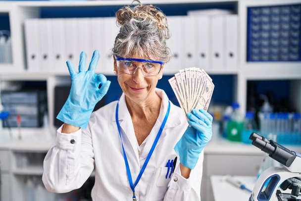 Жінка середнього віку, що працює в науковій лабораторії, тримає долари, роблячи знак ок з пальцями, усміхнений дружній жест відмінний символ
  - Фото, зображення