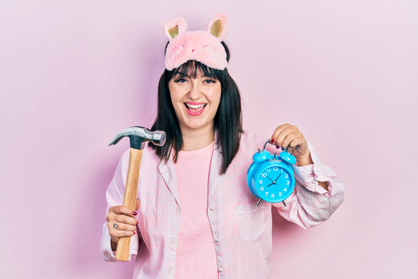 Young hispanic woman wearing sleep mask and robe holding alarm clock and hammer smiling and laughing hard out loud because funny crazy joke.  - 写真・画像