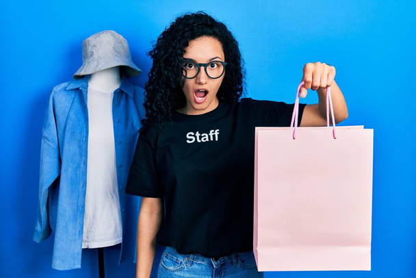 Young hispanic woman with curly hair wearing staff t shirt holding shopping bag scared and amazed with open mouth for surprise, disbelief face  - Foto, imagen
