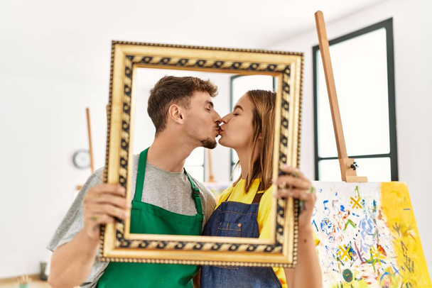 Νέοι Ισπανοί καλλιτέχνες φιλιούνται και κρατούν κενό πλαίσιο στο στούντιο τέχνης. - Φωτογραφία, εικόνα