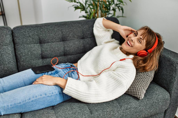 Young caucasian woman listening to music lying on sofa at home - Fotó, kép