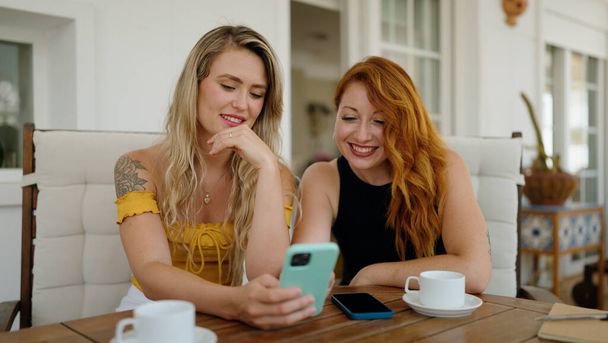 Akıllı telefon kullanan ve kahve içen iki kadın evdeki terasta oturuyor. - Fotoğraf, Görsel
