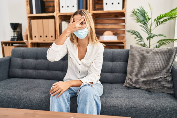 Young blonde woman treating covid fear at therapy peeking in shock covering face and eyes with hand, looking through fingers afraid  - Photo, Image
