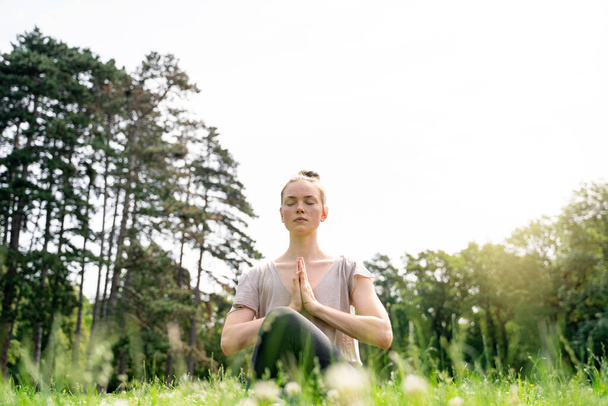 Young caucasian slim fit blond hair carefree calm woman sitting in lotus yoga pose with closed eyes and hands as in prayer, meditating on green grass meadow in summer park on early morning or sunset - Fotó, kép