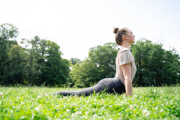 Young caucasian fit woman in light cotton t-shirt doing aerobics, back stretching or sun salutation upward facing dog yoga pose after workout or pilates on grass in summer park on morning or afternoon - Foto, afbeelding