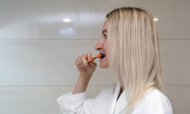 Young blonde woman in a white coat brushes her teeth with a toothbrush and paste, side view. Self-care and daily routine. - Foto, afbeelding