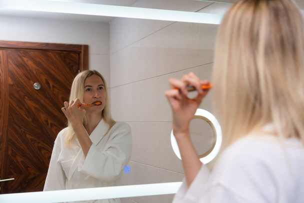 Young blonde woman in a white coat brushes her teeth with a toothbrush and paste, looks in the mirror. Self-care and daily routine. - Foto, immagini
