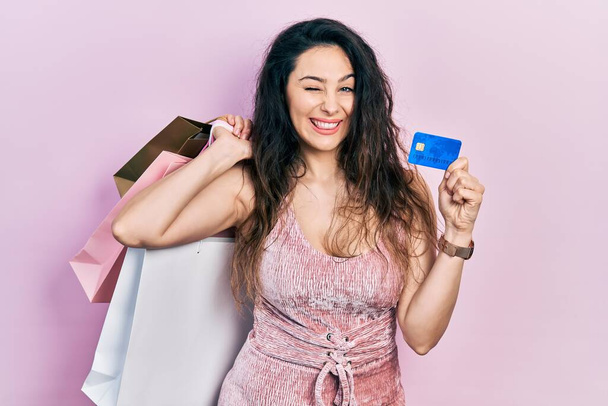 Junge hispanische Frau im Weihnachtsmann-Kostüm mit Einkaufstaschen und Kreditkarte, die augenzwinkernd in die Kamera blickt, mit sexy Ausdruck, fröhlichem und glücklichem Gesicht.  - Foto, Bild