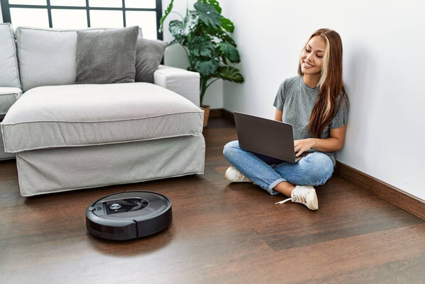Young caucasian girl controlling robot vacuum cleaner with laptop sitting on the floor at home. - Fotografie, Obrázek