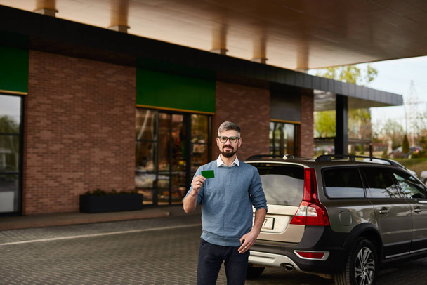 Bearded man in casual clothes demonstrating green plastic card while standing near car at gas station - Valokuva, kuva