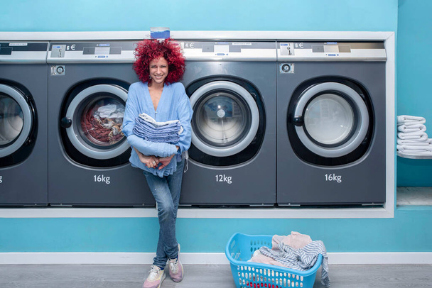 Jeune femme souriante aux cheveux rouges afro tenant des vêtements dans une laverie automatique bleue - Photo, image