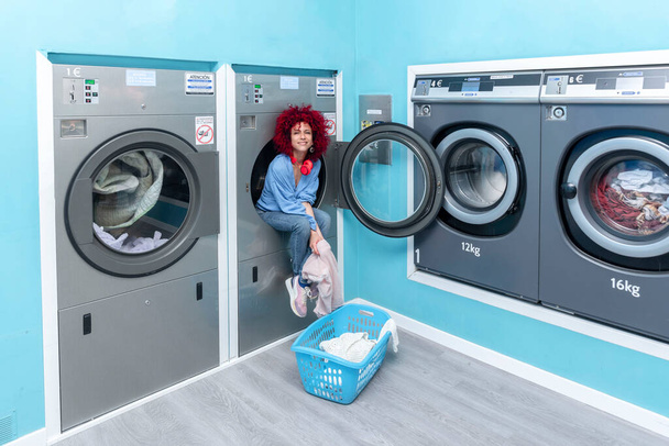Mavi otomatik çamaşır odasında, çamaşır makinesinin içinde oturan, afro saçlı, gülümseyen genç bir Latin kadın. - Fotoğraf, Görsel