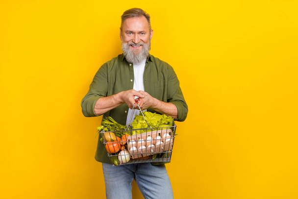 Portrait of positive satisfied aged person toothy smile hold groceries basket isolated on yellow color background. - Foto, imagen