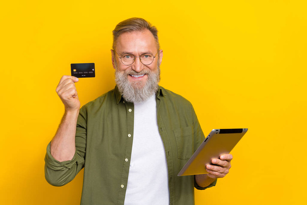 Portrait of positive aged man hold debit plastic card tablet toothy smile isolated on yellow color background. - Foto, afbeelding