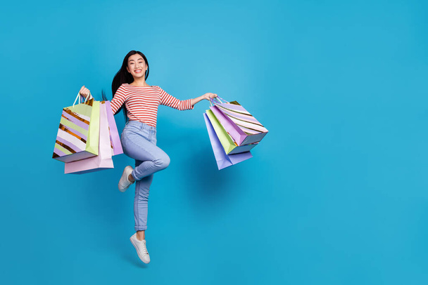 Full length photo of cute funny woman wear striped shirt jumping high holding shoppers empty space isolated blue color background. - 写真・画像