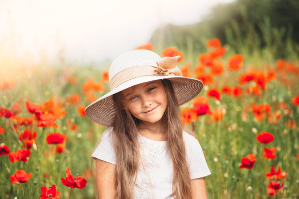 Pequeña chica de pelo largo en sombrero posando en el campo de amapolas con sol de verano - Foto, imagen