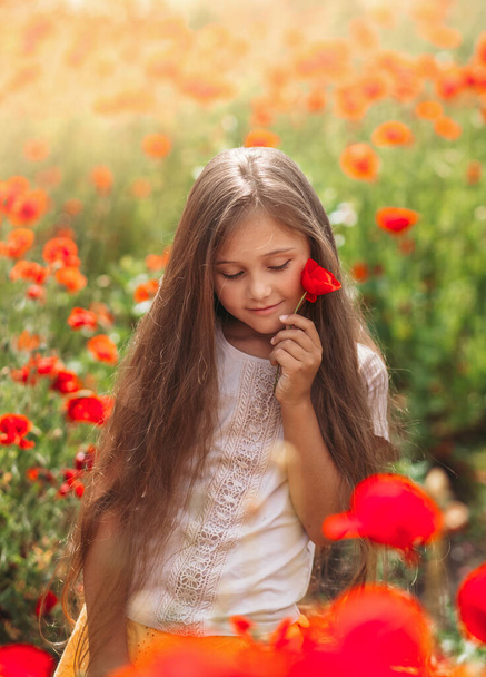 Kleines langhaariges Mädchen posiert auf einem Mohnfeld mit der Sommersonne. Vertikal - Foto, Bild