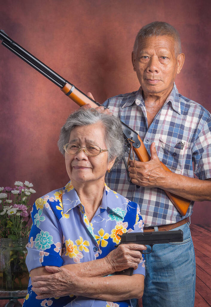 Portré egy idős párról, akik sörétes puskát és sportpuskát tartanak, miközben ülnek, és régi háttérrel állnak. Pneumatikus pisztoly. Az idősek és a sport fogalma. - Fotó, kép