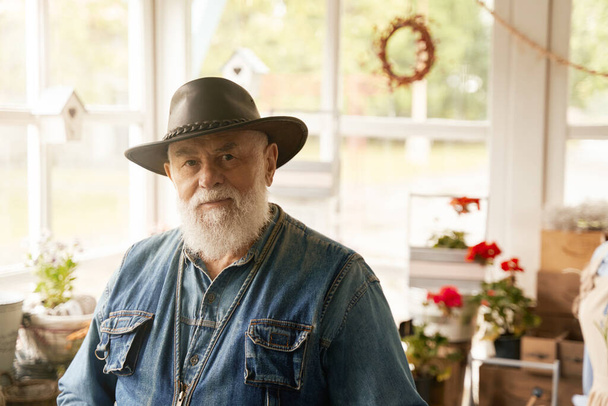 彼の田舎の花屋のブティックの中に立つカウボーイハットのグリズリー高齢者の肖像 - 写真・画像