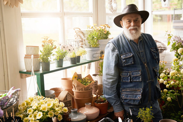 Taille omhoog portret van gelukkige oude bloemist in cowboy hoed werken met potplanten in bloemenwinkel - Foto, afbeelding