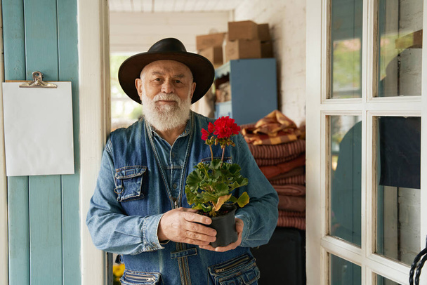 Waist up portrait of smiling senior male holding red geranium while caring about his purchase - Photo, Image