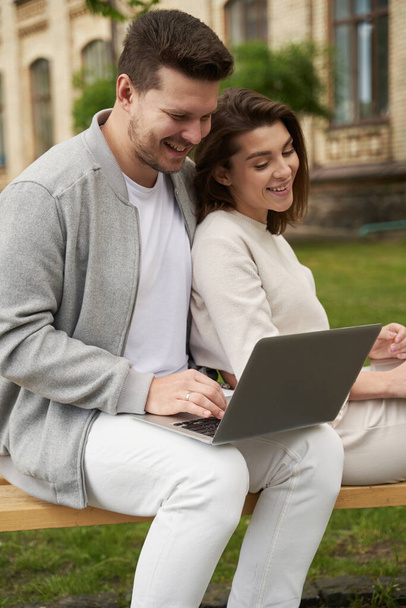 Женщина и влюбленный мужчина сидят на скамейке и улыбаются, смотря видео на открытом воздухе - Фото, изображение