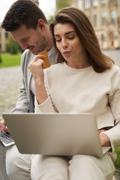 Kobieta trzymająca kartę kredytową banku i patrząca na wyświetlacz laptopa podczas gdy mężczyzna przeglądający internet szuka zakupów na notebooku - Zdjęcie, obraz