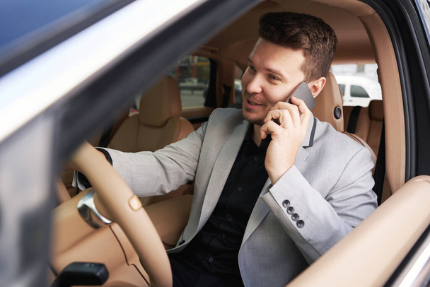 Male entrepreneur having phone conversation and smiling while driving automobile - Photo, Image