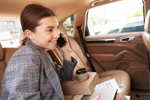 Joyful young woman having phone conversation and smiling while sitting on back seat in automobile - Foto, afbeelding