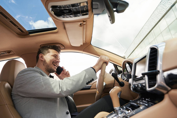 Veselý muž řidič s telefonem konverzace a s úsměvem při řízení auta - Fotografie, Obrázek