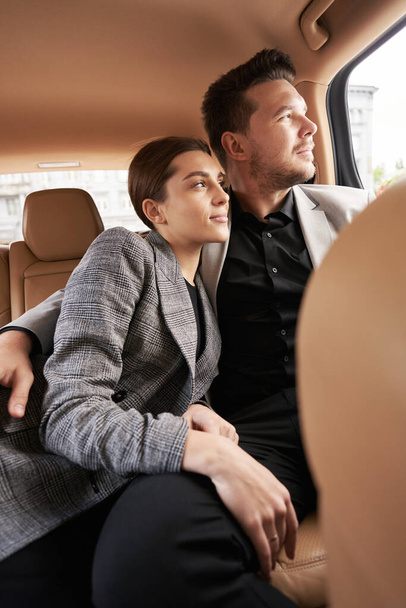 Empresário abraçando mulher e olhando para fora da janela do veículo enquanto sentado no banco de trás no automóvel - Foto, Imagem